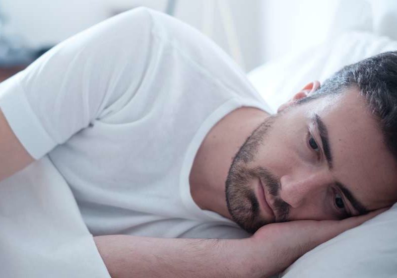 sad man on bed wearing white tshirt Testosterone Therapy How Can It Help, And Is It Safe feat