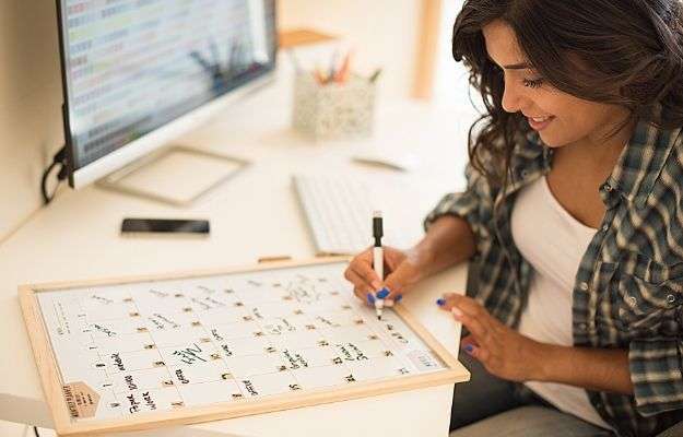 Woman on computer desk writing on a calendar | How to Stay Focused By Planning Your Day