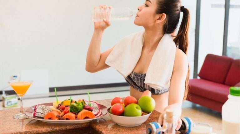 Feature | woman-drinking-water-with-dumbbel-in-her-hand-after-workout-with-salad-and-fruits-on-the-table | 7 Science-Backed Ways To Lose Stubborn Body Fat