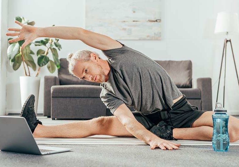 senior man doing stretching on yoga mat while looking at the laptop's screen | feature | What Is A Personalized Weight Loss Program?