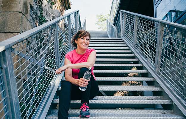 Happy and beautiful middle aged woman sitting on metallic stairs relaxing before running outdoors holding a water bottle energy boost